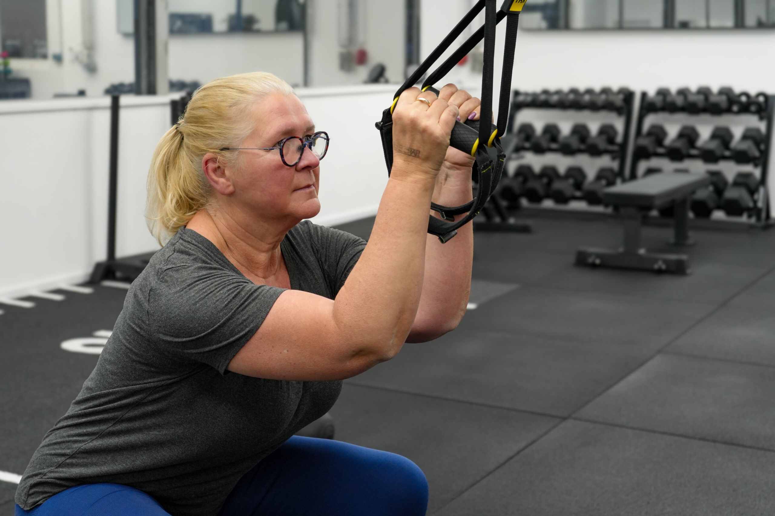Ältere Frau mit blonden Haaren und Brille macht eine Kniebeuge mit TRX-Bändern beim Training in einem modernen Fitnessstudio in Essen, im Hintergrund Hanteln und Trainingsgeräte.