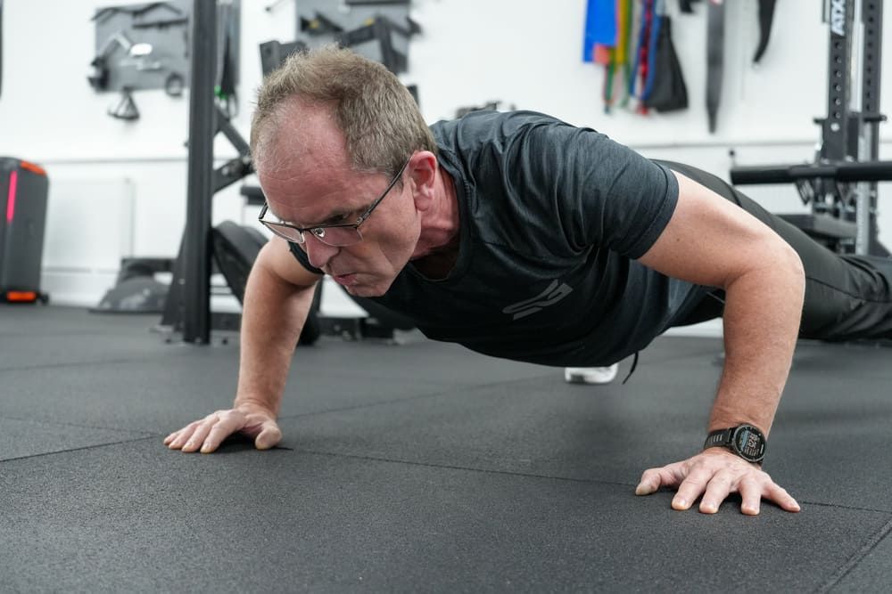 Mann mittleren Alters macht konzentriert Liegestütze auf dem Boden eines modernen Fitnessstudios – effektives Ganzkörpertraining im Bootcamp Essen.