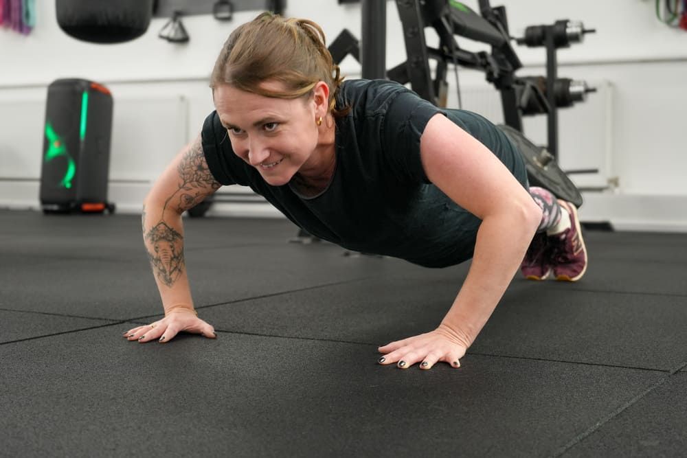 Junge Frau mit Tätowierungen macht motiviert Liegestütze auf dem Boden eines Fitnessstudios, im Hintergrund Fitnessgeräte und Boxsäcke bei dem Bootcamp in Essen
