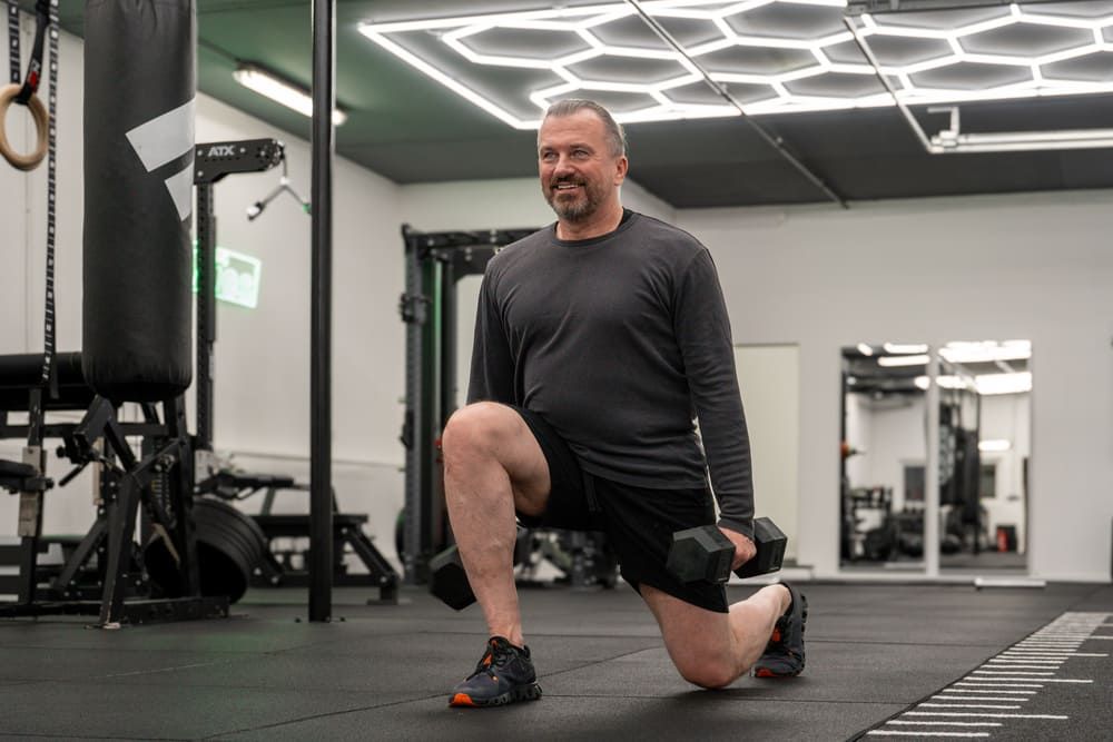 Mann mittleren Alters führt Ausfallschritte mit Kurzhanteln beim Personal Training in einem modernen Fitnessstudio in Essen aus, umgeben von Trainingsgeräten und Boxsäcken.