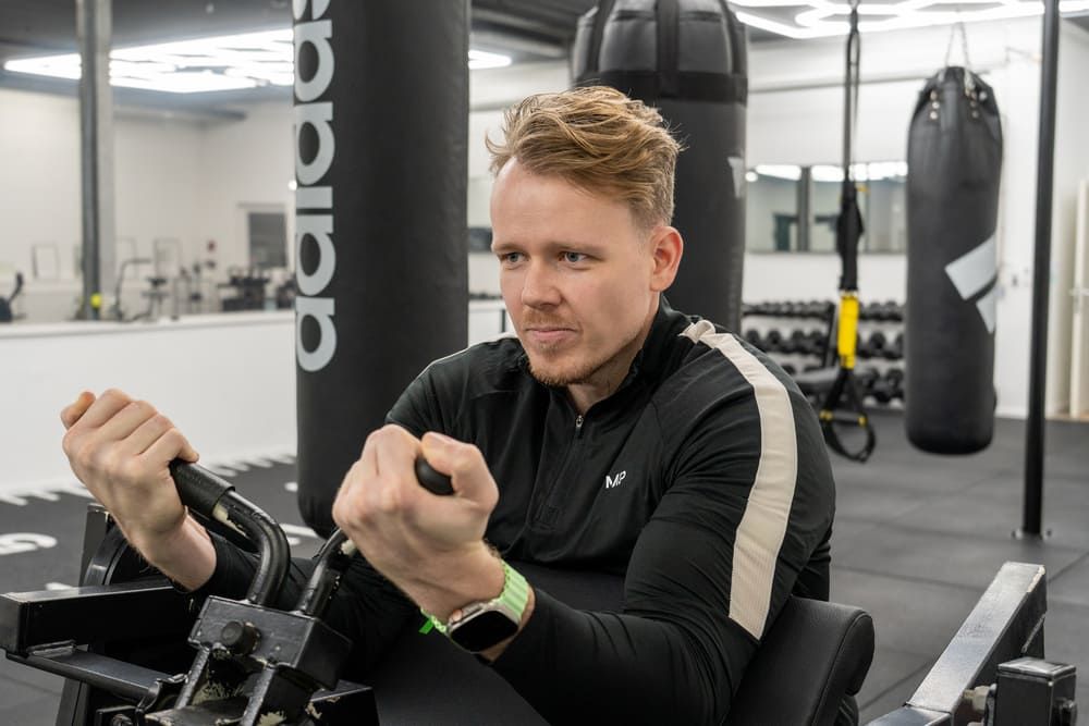 Junger Mann mit sportlicher Kleidung trainiert konzentriert an einem Fitnessgerät in einem modernen Fitnessstudio in Essen, umgeben von Boxsäcken und Hantelbereich.