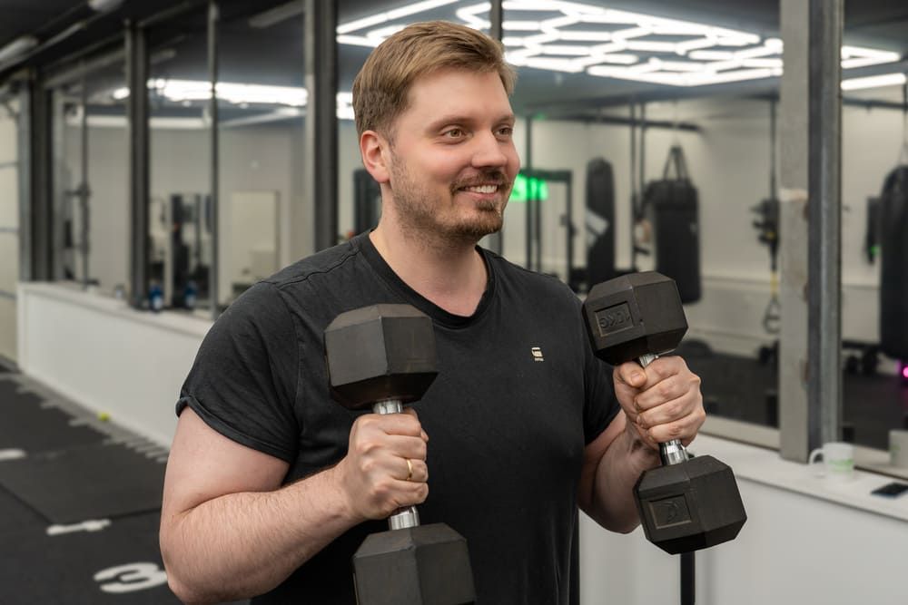 Junger Mann in schwarzem T-Shirt trainiert mit zwei Kurzhanteln in einem modernen Personaltrainingsstudio in Essen, lächelt motiviert während des Trainings.